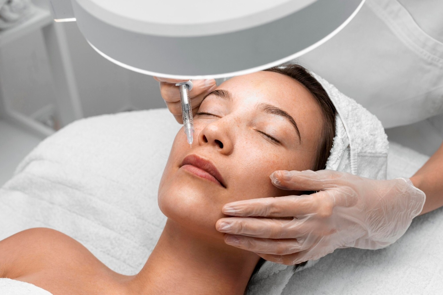 Facial Rejuvenation Richmond KY 1 - The Skin Suite - The Skin Suite Woman lying with eyes closed during a facial; a clinician injects near her eye area using a syringe under bright lamp light.