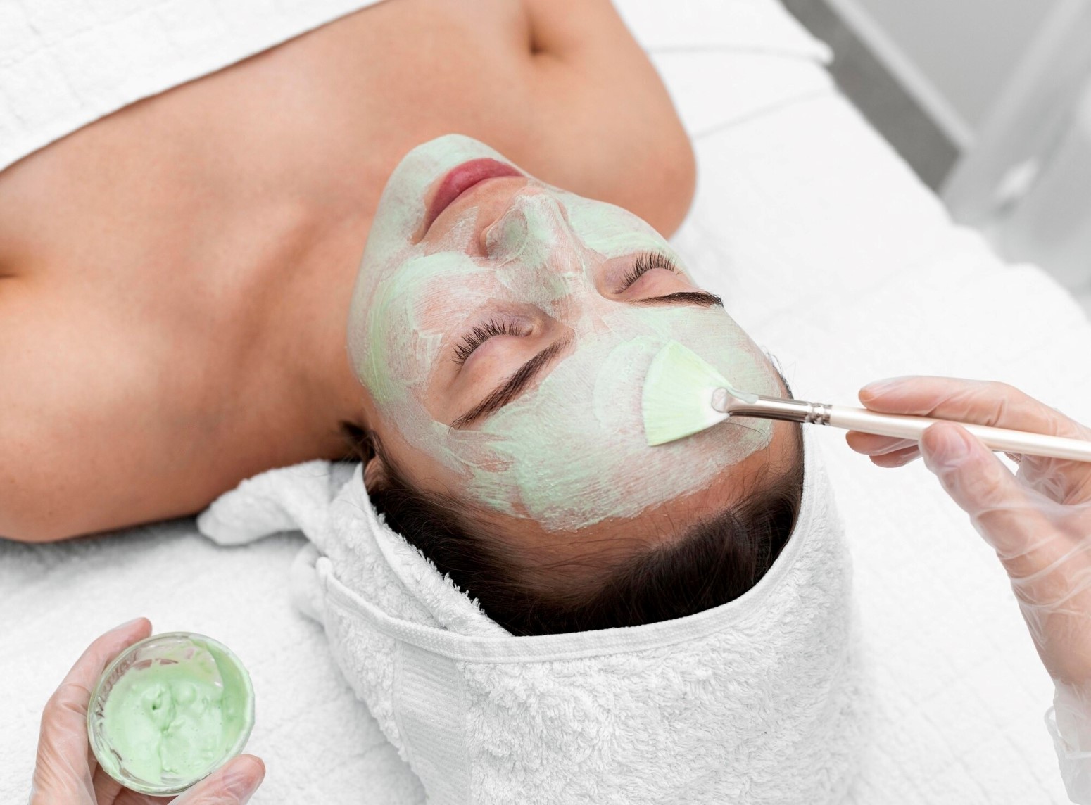 Facial Rejuvenation Richmond KY 2 - The Skin Suite Spa treatment: a woman lies with a mint-green facial mask applied to her face by a practitioner using a brush on her cheek area.