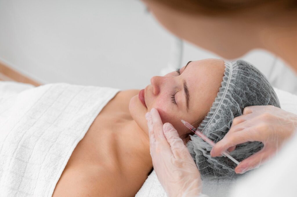 Woman resting on a treatment table as a clinician injects her lips with a syringe, wearing gloves and a hair cap in a clinical setting.