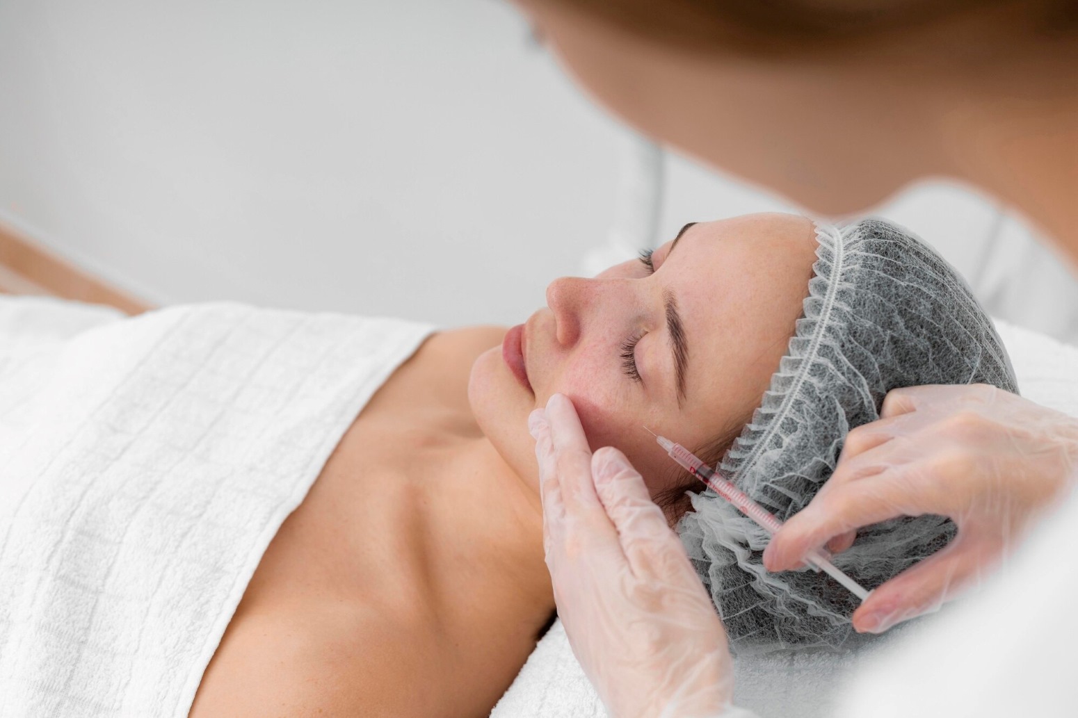 Facial Rejuvenation Richmond KY 3 - The Skin Suite Woman resting on a treatment table as a clinician injects her lips with a syringe, wearing gloves and a hair cap in a clinical setting.