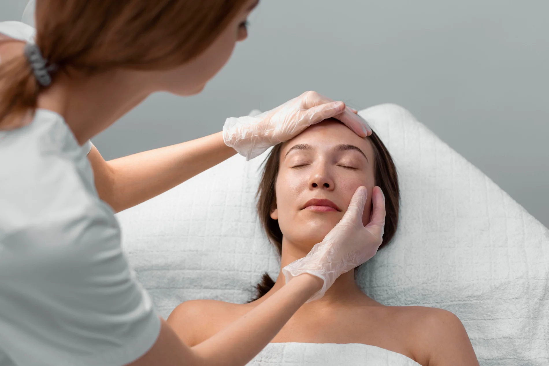 beautician-with-female-client-salon-face-care-routine - The Skin Suite Clinician wearing gloves gently touching a woman's forehead during a facial treatment.
