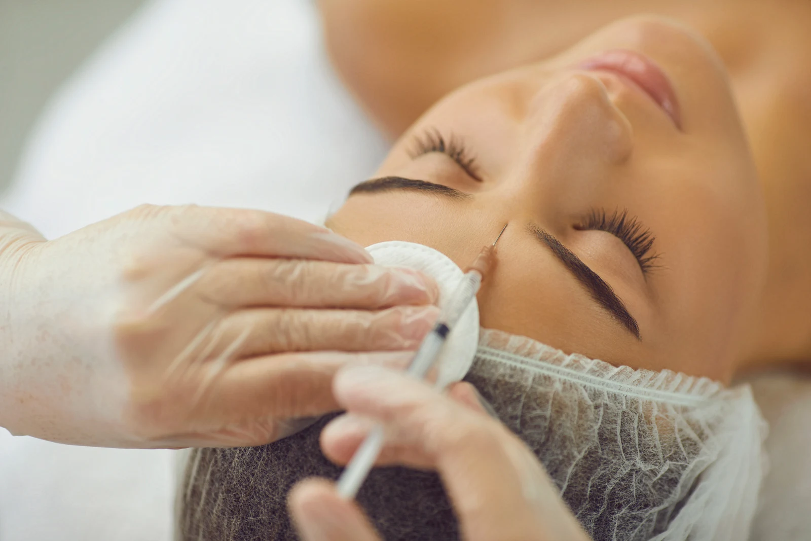 Young woman recieving injection of anti-aging botox filler to forehead - The Skin Suite - The Skin Suite Aesthetician wearing gloves holds a syringe near a clients forehead for a cosmetic injection, with a cotton pad nearby.